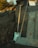 Two wooden-handled gardening tools, a shovel and a rake, are leaning against a concrete wall. Beside them, several wooden planks are stacked. The scene is outdoors, with grass growing near the edges and some trees visible in the blurred background.