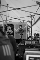 A crowded market stall features a display of posters. The prominent poster depicts a man under the title 'Fight Club'. Metal frames and tarps form an overhead structure suggestive of a typical market setting. Several people are visible browsing in the background.