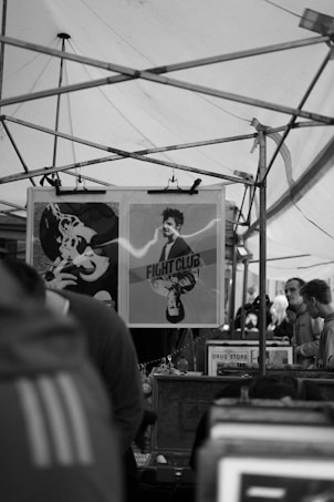 A crowded market stall features a display of posters. The prominent poster depicts a man under the title 'Fight Club'. Metal frames and tarps form an overhead structure suggestive of a typical market setting. Several people are visible browsing in the background.