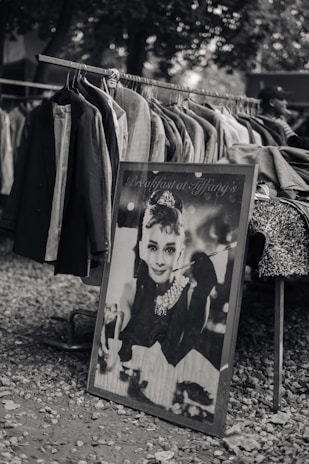A black and white photograph of a market stall displaying a rack filled with hanging coats and jackets. Propped against the rack is a framed poster featuring an elegant woman with a pearl necklace and an iconic hairstyle, holding a cigarette holder. The setting is outdoors with a backdrop of trees, and the ground is covered with small stones.