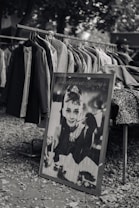 A black and white photograph of a market stall displaying a rack filled with hanging coats and jackets. Propped against the rack is a framed poster featuring an elegant woman with a pearl necklace and an iconic hairstyle, holding a cigarette holder. The setting is outdoors with a backdrop of trees, and the ground is covered with small stones.