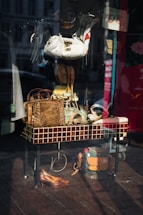 The image captures a storefront display with various accessories and items arranged on and around a tiled table. Visible objects include handbags made of different materials, a pair of shoes, hats, and belts. The reflection on the glass shows a street scene with buildings and vehicles.