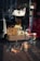 The image captures a storefront display with various accessories and items arranged on and around a tiled table. Visible objects include handbags made of different materials, a pair of shoes, hats, and belts. The reflection on the glass shows a street scene with buildings and vehicles.