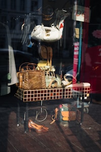 The image captures a storefront display with various accessories and items arranged on and around a tiled table. Visible objects include handbags made of different materials, a pair of shoes, hats, and belts. The reflection on the glass shows a street scene with buildings and vehicles.