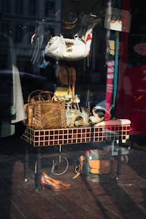 The image captures a storefront display with various accessories and items arranged on and around a tiled table. Visible objects include handbags made of different materials, a pair of shoes, hats, and belts. The reflection on the glass shows a street scene with buildings and vehicles.