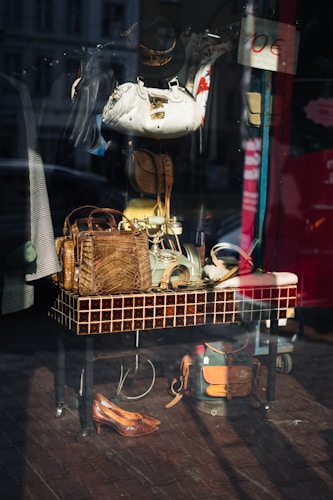 The image captures a storefront display with various accessories and items arranged on and around a tiled table. Visible objects include handbags made of different materials, a pair of shoes, hats, and belts. The reflection on the glass shows a street scene with buildings and vehicles.