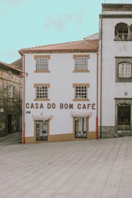 A quaint building with white walls and brown trim features the words 'Casa do Bom Café' in large letters on the front. It is situated in a quiet, partially paved square and is flanked by other traditional stone buildings.