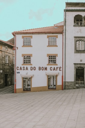 A quaint building with white walls and brown trim features the words 'Casa do Bom Café' in large letters on the front. It is situated in a quiet, partially paved square and is flanked by other traditional stone buildings.