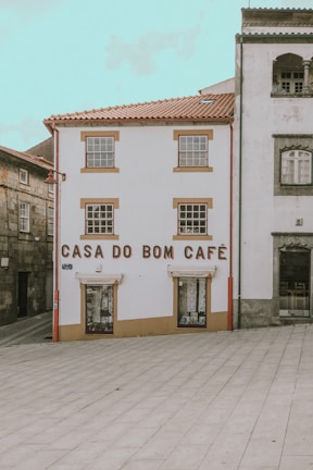 A quaint building with white walls and brown trim features the words 'Casa do Bom Café' in large letters on the front. It is situated in a quiet, partially paved square and is flanked by other traditional stone buildings.