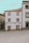 A quaint building with white walls and brown trim features the words 'Casa do Bom Café' in large letters on the front. It is situated in a quiet, partially paved square and is flanked by other traditional stone buildings.