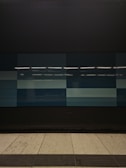 A tiled wall featuring a series of rectangular panels in varying shades of blue, situated in what appears to be an indoor setting. The tiles are clean and sleek, with overhead lighting reflecting off the surface, creating a geometric and modern appearance. The floor in front is tiled in light gray with dark trim along the edge.