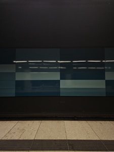 A tiled wall featuring a series of rectangular panels in varying shades of blue, situated in what appears to be an indoor setting. The tiles are clean and sleek, with overhead lighting reflecting off the surface, creating a geometric and modern appearance. The floor in front is tiled in light gray with dark trim along the edge.