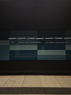 A tiled wall featuring a series of rectangular panels in varying shades of blue, situated in what appears to be an indoor setting. The tiles are clean and sleek, with overhead lighting reflecting off the surface, creating a geometric and modern appearance. The floor in front is tiled in light gray with dark trim along the edge.