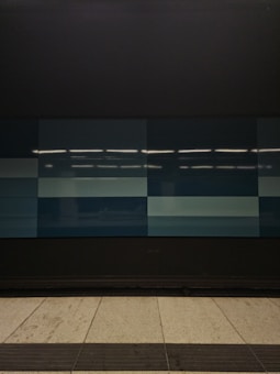 A tiled wall featuring a series of rectangular panels in varying shades of blue, situated in what appears to be an indoor setting. The tiles are clean and sleek, with overhead lighting reflecting off the surface, creating a geometric and modern appearance. The floor in front is tiled in light gray with dark trim along the edge.