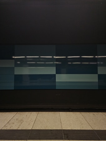 A tiled wall featuring a series of rectangular panels in varying shades of blue, situated in what appears to be an indoor setting. The tiles are clean and sleek, with overhead lighting reflecting off the surface, creating a geometric and modern appearance. The floor in front is tiled in light gray with dark trim along the edge.