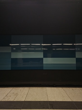 A tiled wall featuring a series of rectangular panels in varying shades of blue, situated in what appears to be an indoor setting. The tiles are clean and sleek, with overhead lighting reflecting off the surface, creating a geometric and modern appearance. The floor in front is tiled in light gray with dark trim along the edge.