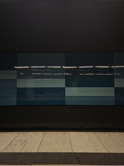 A tiled wall featuring a series of rectangular panels in varying shades of blue, situated in what appears to be an indoor setting. The tiles are clean and sleek, with overhead lighting reflecting off the surface, creating a geometric and modern appearance. The floor in front is tiled in light gray with dark trim along the edge.