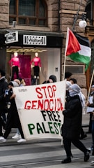 A group of people participate in a street protest. They hold a large banner that reads 'Stop Genocide Free Palestine'. One of the protestors is carrying a flag that represents Palestine. The scene is set in front of a storefront with mannequins wearing bright pink tops.