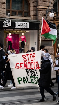 A group of people participate in a street protest. They hold a large banner that reads 'Stop Genocide Free Palestine'. One of the protestors is carrying a flag that represents Palestine. The scene is set in front of a storefront with mannequins wearing bright pink tops.