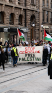 A group of people are marching on a city street holding a large banner with the words 'STOP GENOCIDE FREE PALESTINE'. Several individuals are carrying Palestinian flags. The setting appears to be an urban area lined with stone buildings and shops.