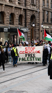 A group of people are marching on a city street holding a large banner with the words 'STOP GENOCIDE FREE PALESTINE'. Several individuals are carrying Palestinian flags. The setting appears to be an urban area lined with stone buildings and shops.