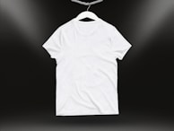 A plain white t-shirt on a hanger against a dark background. Soft lighting highlights the texture and folds of the fabric.