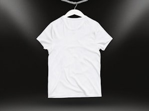 A plain white t-shirt on a hanger against a dark background. Soft lighting highlights the texture and folds of the fabric.