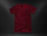 Flat lay of folded Nari t-shirts in warm stone beige and deep red accent colors on volcanic stone surface.