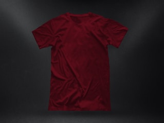 A plain, deep red t-shirt placed against a dark textured background. The garment appears to be crumpled slightly, showing natural folds and shadows. The fabric looks soft, and the shirt is displayed in a casual, relaxed style.