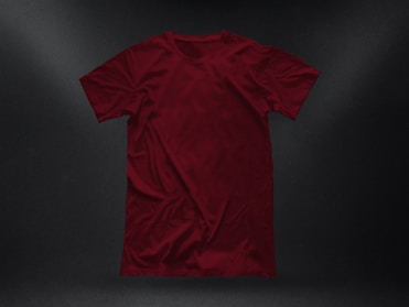 A plain, deep red t-shirt placed against a dark textured background. The garment appears to be crumpled slightly, showing natural folds and shadows. The fabric looks soft, and the shirt is displayed in a casual, relaxed style.
