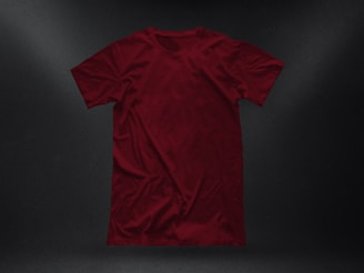 A plain, deep red t-shirt placed against a dark textured background. The garment appears to be crumpled slightly, showing natural folds and shadows. The fabric looks soft, and the shirt is displayed in a casual, relaxed style.
