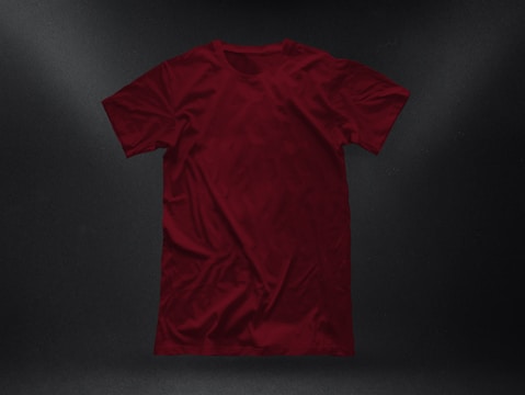 A plain, deep red t-shirt placed against a dark textured background. The garment appears to be crumpled slightly, showing natural folds and shadows. The fabric looks soft, and the shirt is displayed in a casual, relaxed style.
