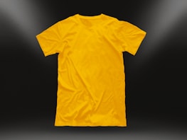 A plain yellow t-shirt is centered against a dark background, illuminated by subtle light gradients in the corners. The shirt is neatly laid out, highlighting its texture and smooth fabric.