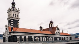 An architectural structure with a distinctive tower and a series of arched windows is captured. The building features dark stone walls contrasted by white accents and red tiled roofing. Ornate designs and decorative elements are present around the windows and along the rooftop areas.