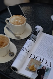 On a glass-topped table, there are two cups of cappuccino, each placed on a saucer with a spoon. Next to the cups, a smartphone is lying flat. A magazine with images and text is partially rolled, displaying black and white photography as well as an article with large bold letters.