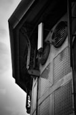 A black and white image of an industrial building facade featuring large ventilation ducts and glass block windows. The structure appears to be metal and concrete, with various pipes and beams integrated into the design. The angle suggests looking upwards at the side of the building.