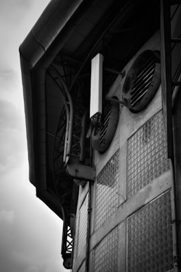 A black and white image of an industrial building facade featuring large ventilation ducts and glass block windows. The structure appears to be metal and concrete, with various pipes and beams integrated into the design. The angle suggests looking upwards at the side of the building.