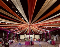 A vibrant and festive outdoor setting with colorful draped fabric radiating from a central point on the ceiling, creating a tent-like structure illuminated by strings of lights. Chairs are arranged in lines, and people are seated with some walking around. The environment conveys a celebratory atmosphere.