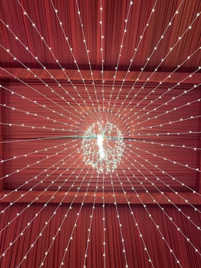 A ceiling decorated with a radiant array of string lights converging to a central chandelier. The backdrop is a richly colored maroon fabric that enhances the warm glow of the lights, creating a starburst pattern toward the chandelier.
