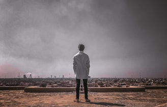 An individual standing on a rooftop wearing a zidd jacket, looking out over the city skyline with determination.