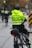 A police officer wearing a bright yellow jacket with 'Vancouver Police' written on it is riding a bike on a city street. The officer is part of a group of police officers on bicycles, and they are heading down a wet pavement, indicating recent rain. The background shows traffic lights, street indicators, and autumn foliage.