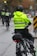 A police officer wearing a bright yellow jacket with 'Vancouver Police' written on it is riding a bike on a city street. The officer is part of a group of police officers on bicycles, and they are heading down a wet pavement, indicating recent rain. The background shows traffic lights, street indicators, and autumn foliage.