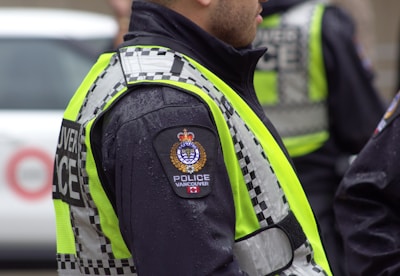 A person wearing a police uniform with a high-visibility vest that features the word 'POLICE VANCOUVER' and an emblem. The vest has a checkered pattern, and the uniform appears to be wet from rain. The background includes a blurred vehicle.
