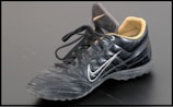 A single black leather sports shoe, featuring a swoosh design on its side. The shoe has black laces loosely tied and a beige inner lining visible at the ankle. It appears slightly worn, with dirt marks on the sole and creases in the leather.