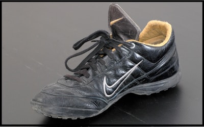 A single black leather sports shoe, featuring a swoosh design on its side. The shoe has black laces loosely tied and a beige inner lining visible at the ankle. It appears slightly worn, with dirt marks on the sole and creases in the leather.