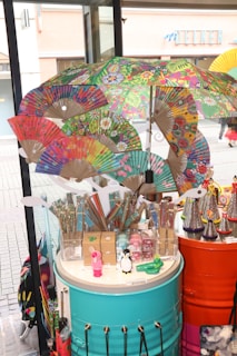 A vibrant display of colorful paper fans, umbrellas, and assorted novelty items on cylindrical tables in a shop or market setting. The fans and umbrellas feature intricate, multicolored designs with floral and abstract patterns. Various small toys and trinkets, including figurines of a penguin and a rabbit, are also visible. The display is set against a backdrop of large glass windows showing a street scene outside.