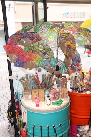 A vibrant display of colorful paper fans, umbrellas, and assorted novelty items on cylindrical tables in a shop or market setting. The fans and umbrellas feature intricate, multicolored designs with floral and abstract patterns. Various small toys and trinkets, including figurines of a penguin and a rabbit, are also visible. The display is set against a backdrop of large glass windows showing a street scene outside.