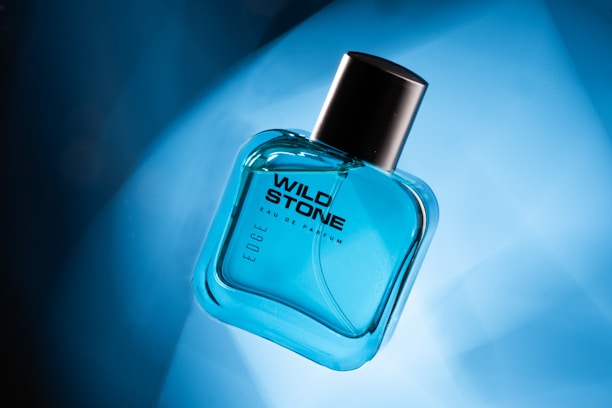 A transparent, blue-tinted glass bottle of Wild Stone Eau de Parfum is positioned at an angle on a gradient blue background. The bottle has a black cap and features modern, bold typography.