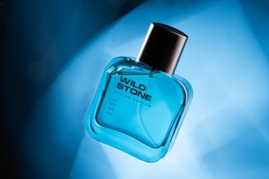 A transparent, blue-tinted glass bottle of Wild Stone Eau de Parfum is positioned at an angle on a gradient blue background. The bottle has a black cap and features modern, bold typography.