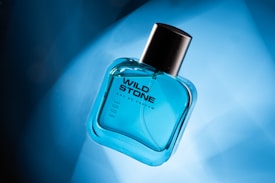 A transparent, blue-tinted glass bottle of Wild Stone Eau de Parfum is positioned at an angle on a gradient blue background. The bottle has a black cap and features modern, bold typography.